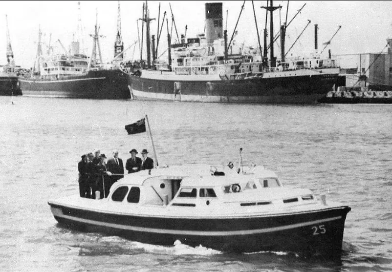 Archival black-and-white photograph of the launch carrying suited officials past cargo ships in the Port of Melbourne